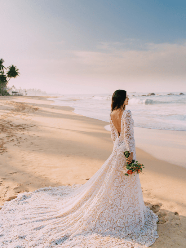 Bride in long lace wedding gown stands on the beach watching the waves
