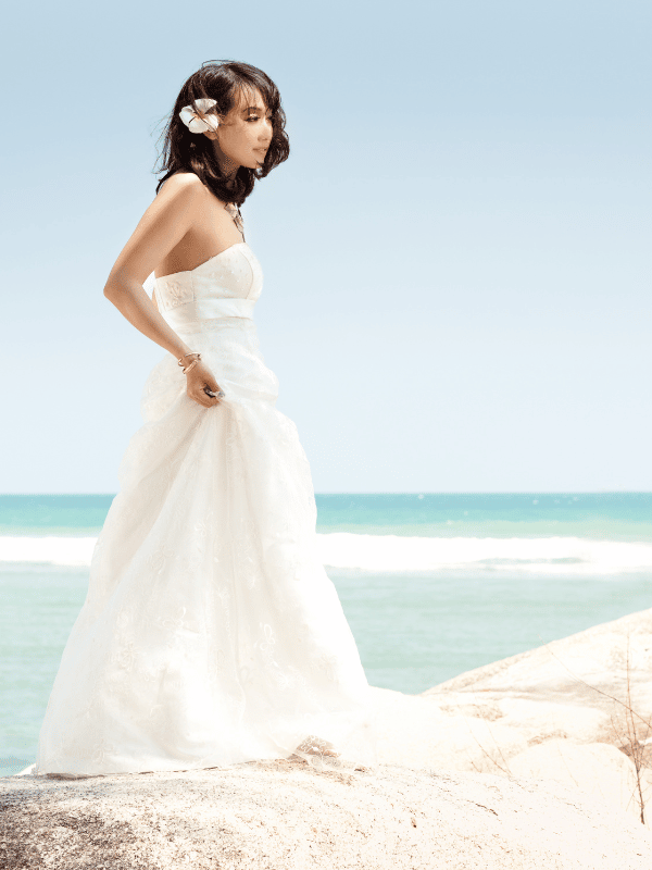 Bride in long white gown stands next to the ocean with a flower in her hair