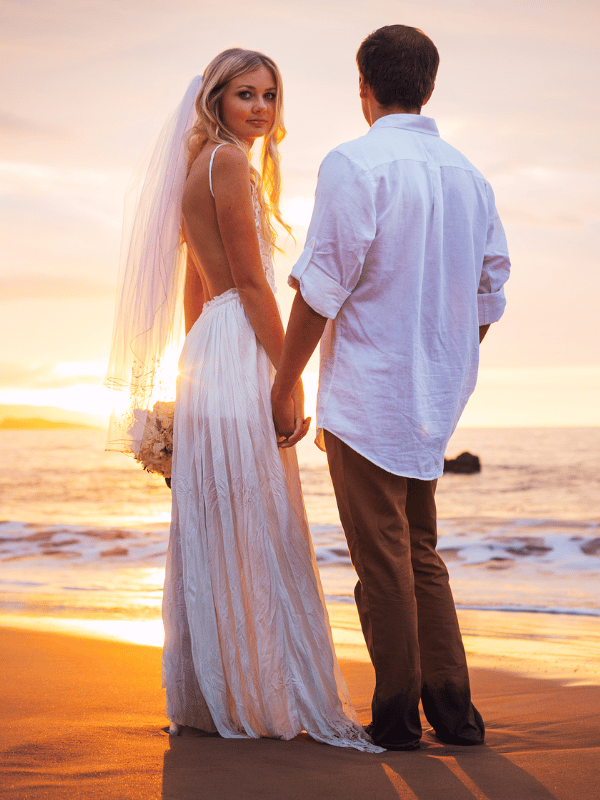 A bride and groom hold hands at sunset on the beach as the bride looks over her shoulder