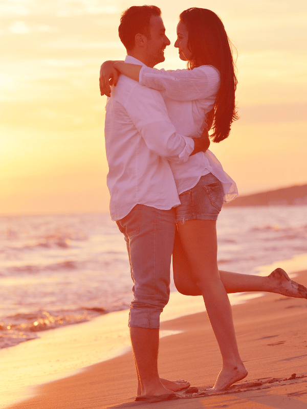 A man and a woman hug on the beach during sunset and the woman lifts her leg behind her
