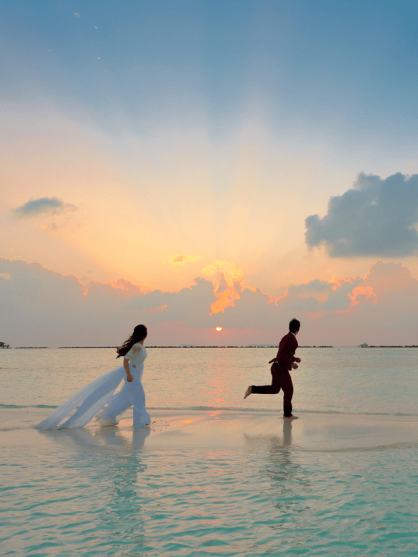 Bride and groom run through shallow ocean waters as the sun sets in the background