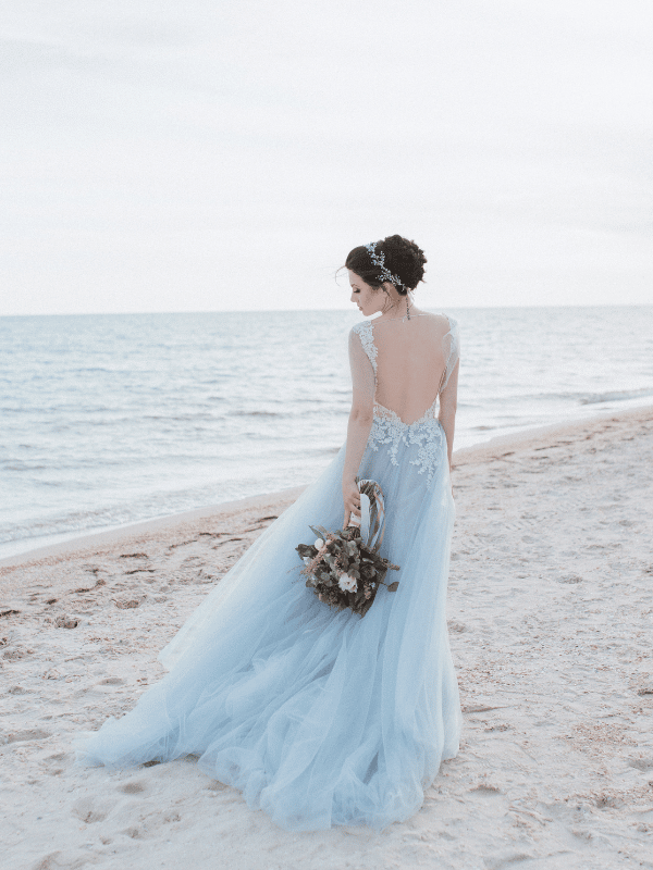 Bride in a blue dress stands on the beach holding a bouquet