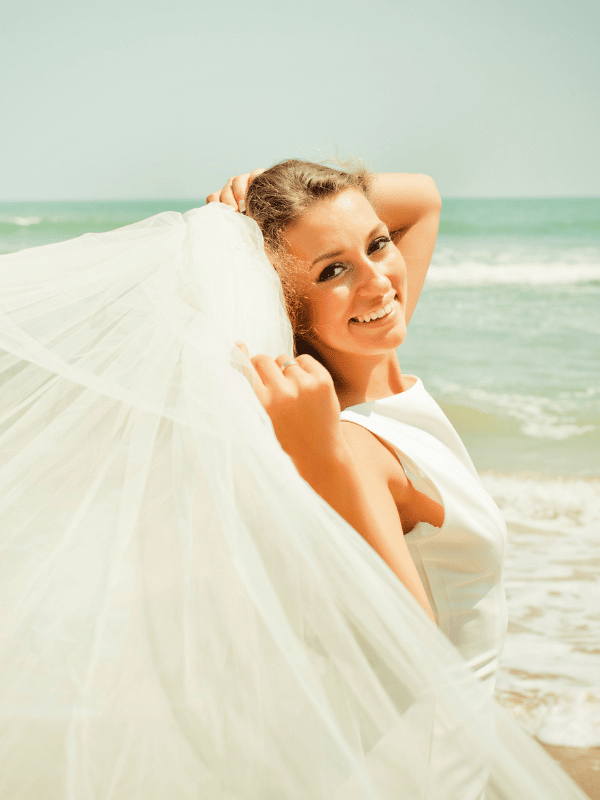 Bride smiles and holds veil as it blows on the wind on the beach