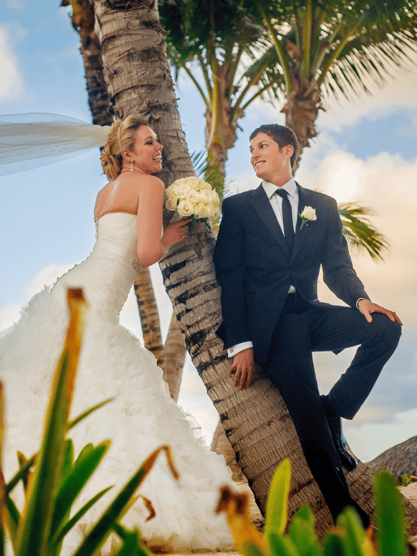 Bride and Groom lean on a palm tree while the bride holds a bouquet of white flowers
