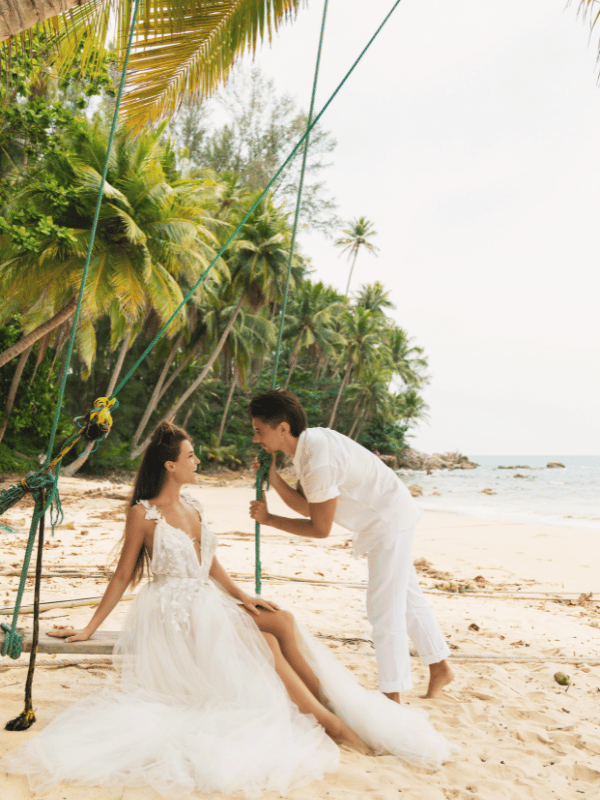 Bride sits on swing on a tropical beach while her groom holds one of the ropes