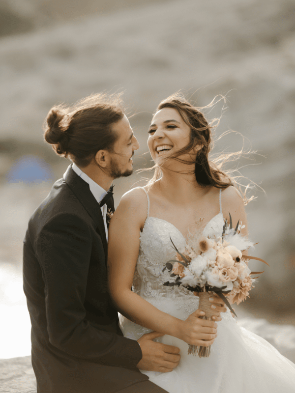 Bride holding bouquet and groom smile at each other on the beach