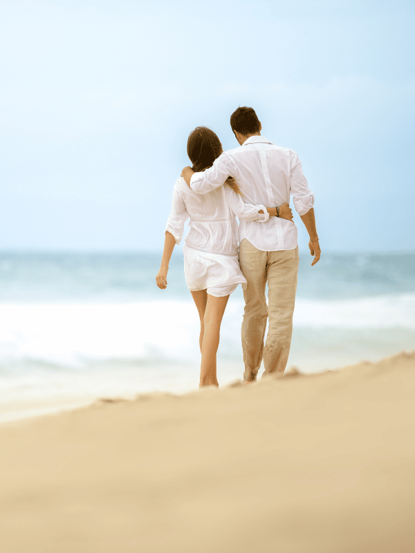 A man and a woman in white walk on the beach with their arms around each other