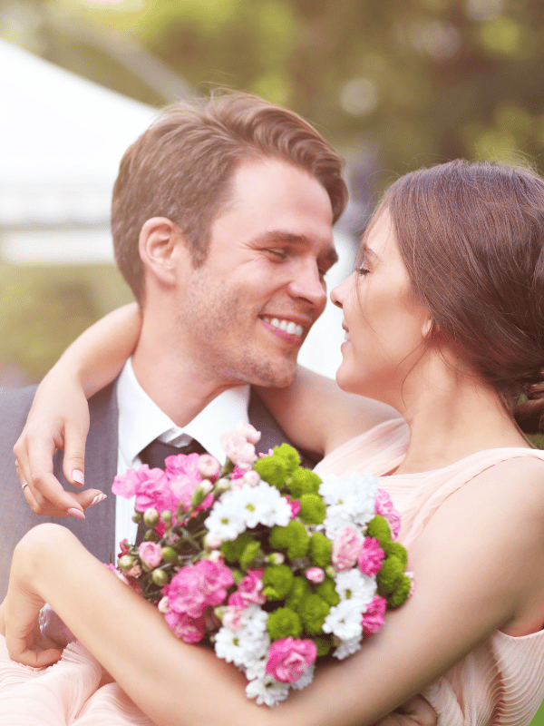 A bride holds a bouquet in one hand and wraps the other arm around her groom's shoulders as they smile at each other