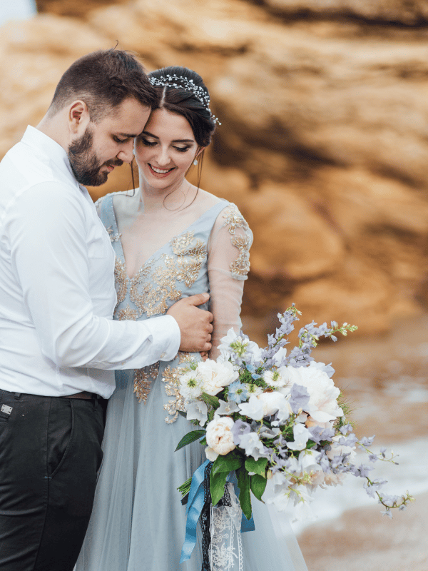 Groom embraces bride in a blue dress with flowers next to a rock formation on the beach