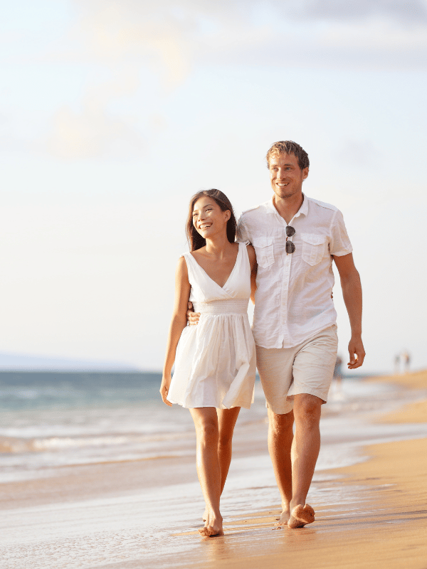 A man and woman wearing white walk along the beach together