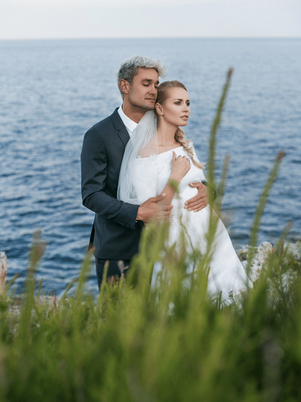 A bride and groom stand in front of the ocean with grass in the foreground