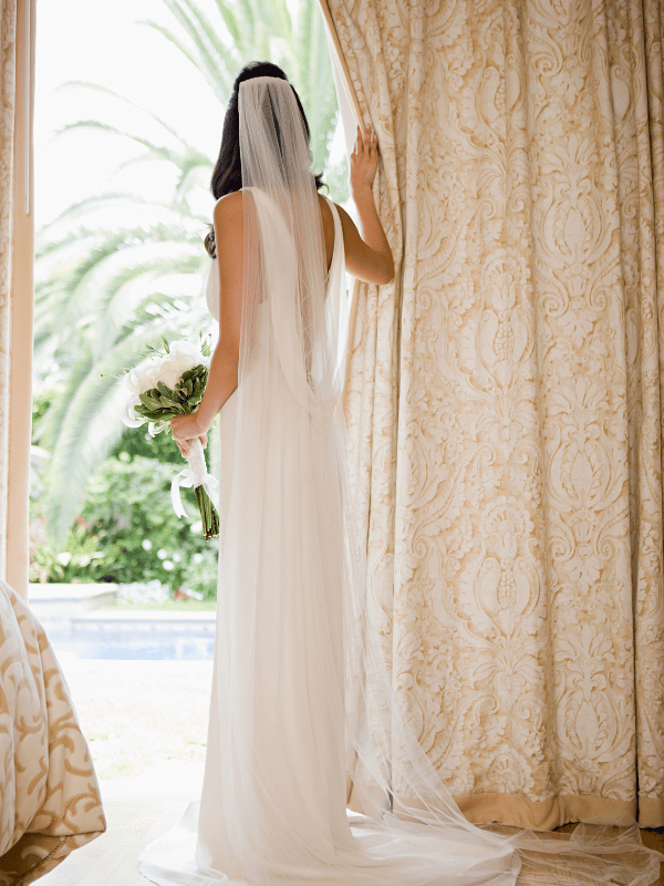 A bride in a wedding dress and veil pulls back a curtain to look at the tropical setting outside