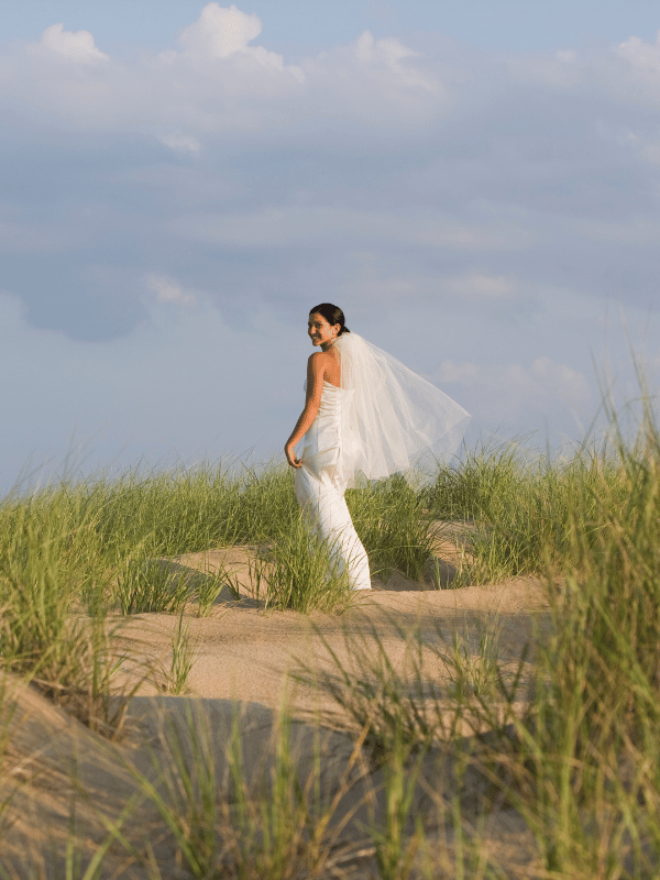 Bride stands on a grass-covered beach as her veil blows in the wind
