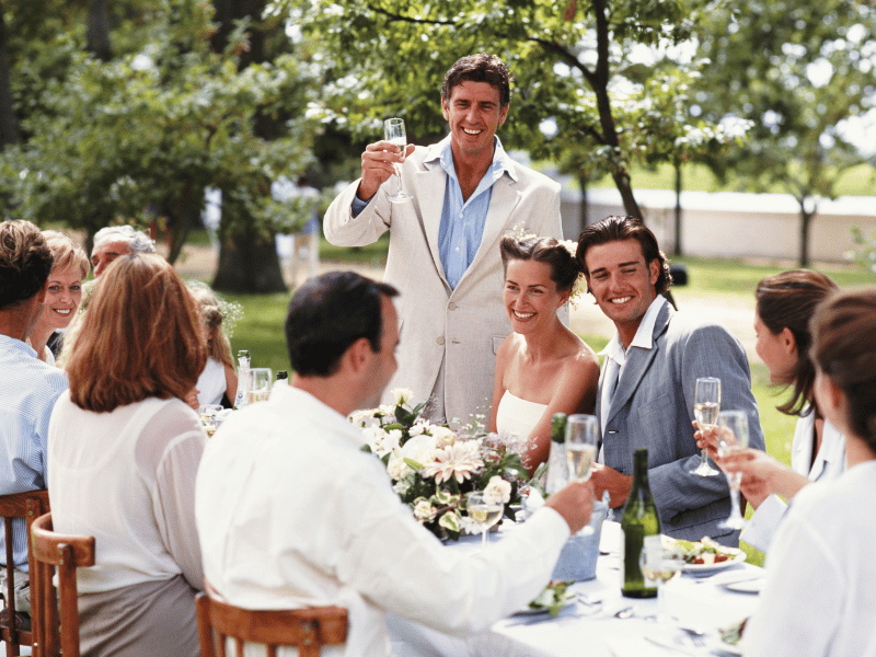 Wedding guests in blue and white sit at a reception table as the best man toasts the couple