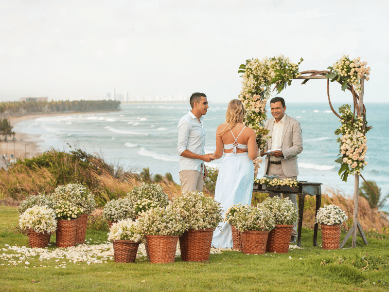 A wedding couple stands with an officiant by the sea surrounded by baskets of flowers.