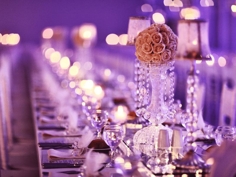 Purple-lit wedding reception tables with candles, pink roses, and crystal decorations.
