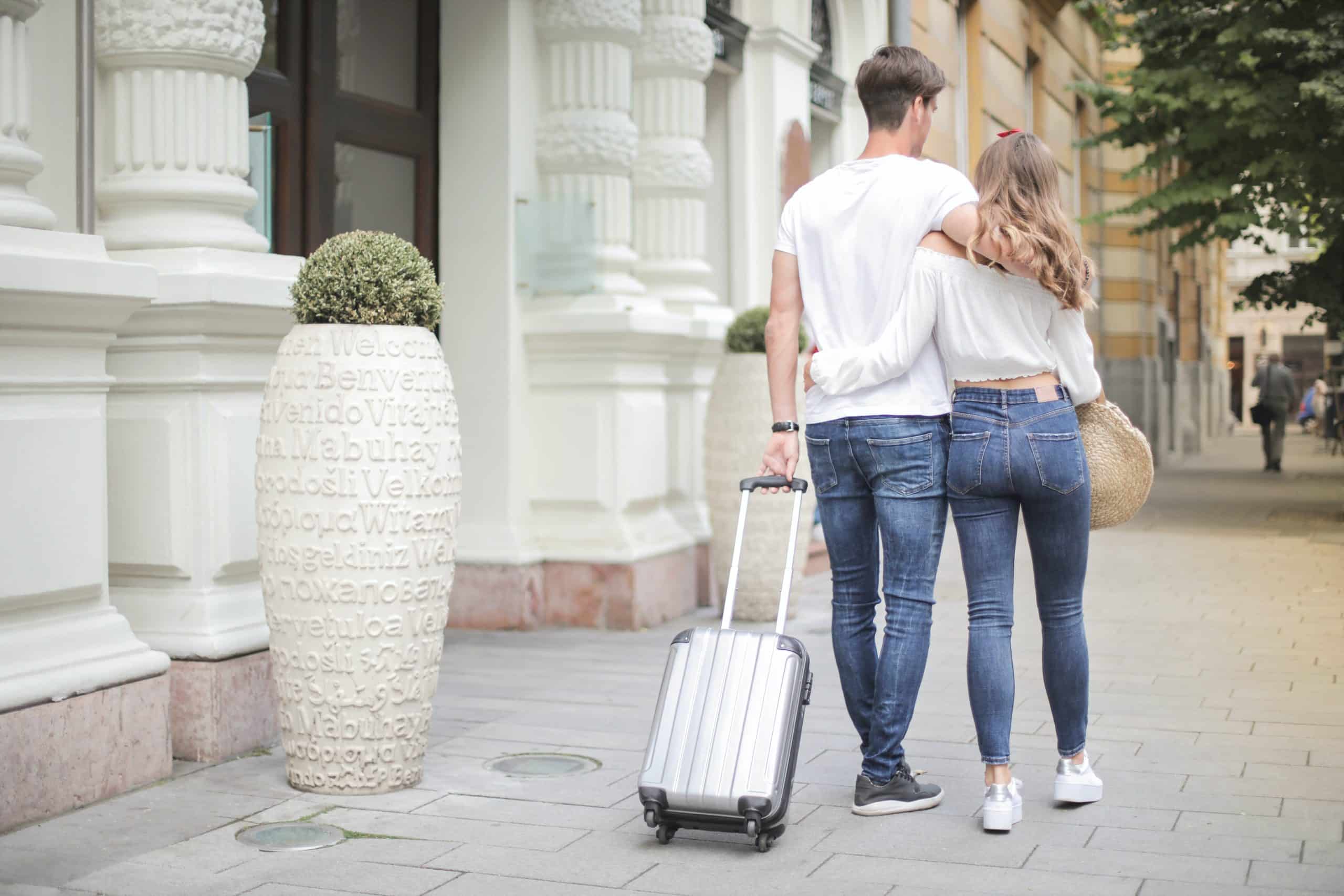 Man and woman with suitcase wrap arms around each other while walking down a city street.