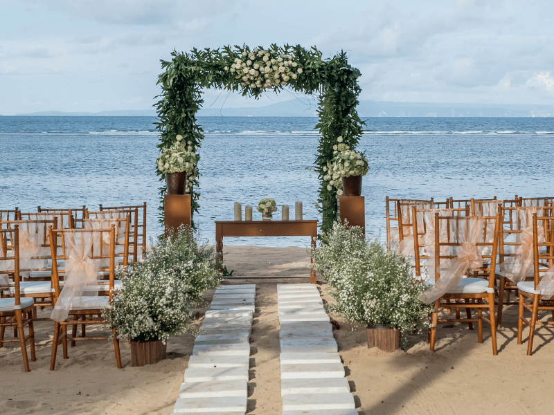 Wedding ceremony setup on the beach with greenery and wooden chairs.