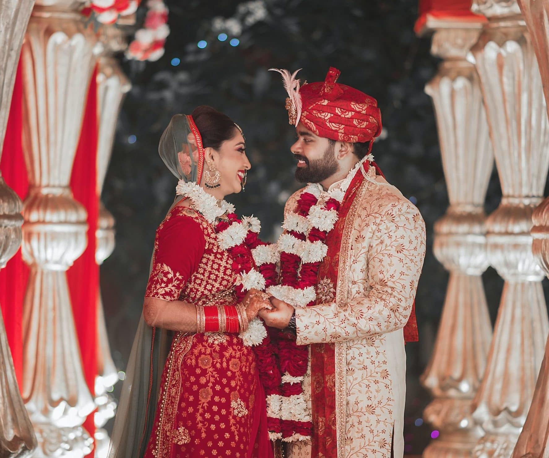 Indian couple in wedding clothes and floral har stand in front of a red and gold backdrop and hold hands.