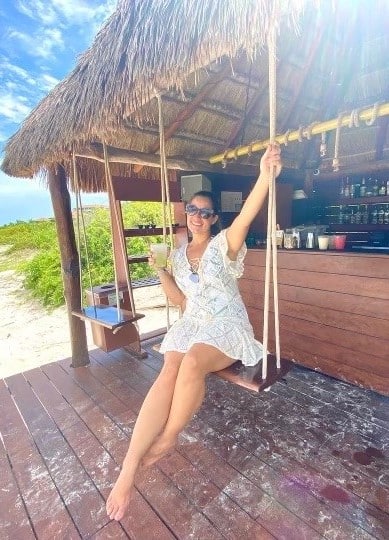 Freelance writer for Destination Brides Kara Adamo sits on a swing at a tiki bar with a tropical drink in her hand
