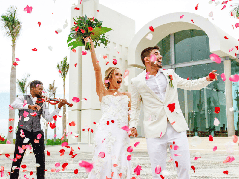 Newlywed couple exits a chapel showered with rose petals while a violinist plays in the background
